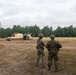 Two soldiers in camouflage uniforms and helmets stand in a dirt field observing a tracked military vehicle, likely an M109 Paladin self-propelled howitzer or similar armored artillery platform. The scene appears to be a training exercise at a military installation, with a treeline of mixed conifers and deciduous trees forming the background. The overcast sky and muddy terrain suggest recent or ongoing field operations.