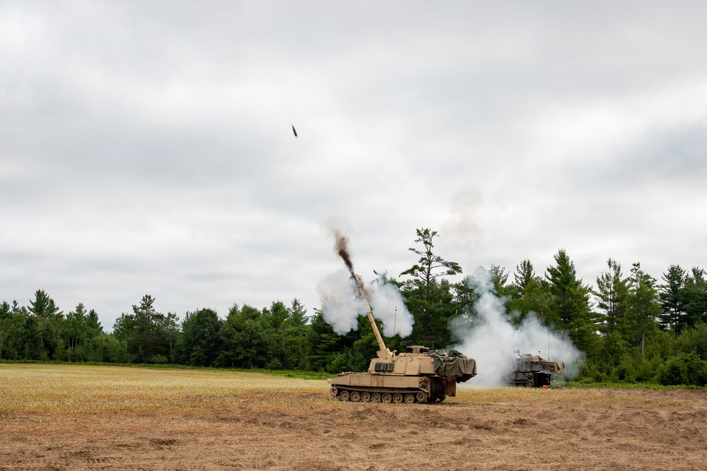 A self-propelled artillery piece, consistent with an M109 Paladin howitzer, is captured mid-fire in an open field with a projectile visibly in flight above the barrel. White and black smoke billows from the muzzle and surrounds the vehicle. A second military vehicle is visible to the right in the background.