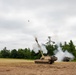A self-propelled artillery piece, consistent with an M109 Paladin howitzer, is captured mid-fire in an open field with a projectile visibly in flight above the barrel. White and black smoke billows from the muzzle and surrounds the vehicle. A second military vehicle is visible to the right in the background.