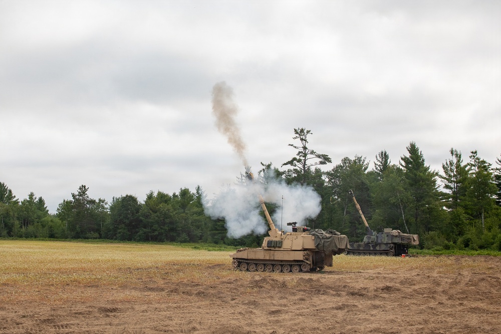 Two self-propelled artillery vehicles are positioned in an open dirt field with a treeline in the background. The foreground vehicle has just fired its main gun, producing a large plume of white and brown smoke rising into an overcast sky. This appears to be a military training exercise or live-fire demonstration.