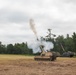 Two self-propelled artillery vehicles are positioned in an open dirt field with a treeline in the background. The foreground vehicle has just fired its main gun, producing a large plume of white and brown smoke rising into an overcast sky. This appears to be a military training exercise or live-fire demonstration.