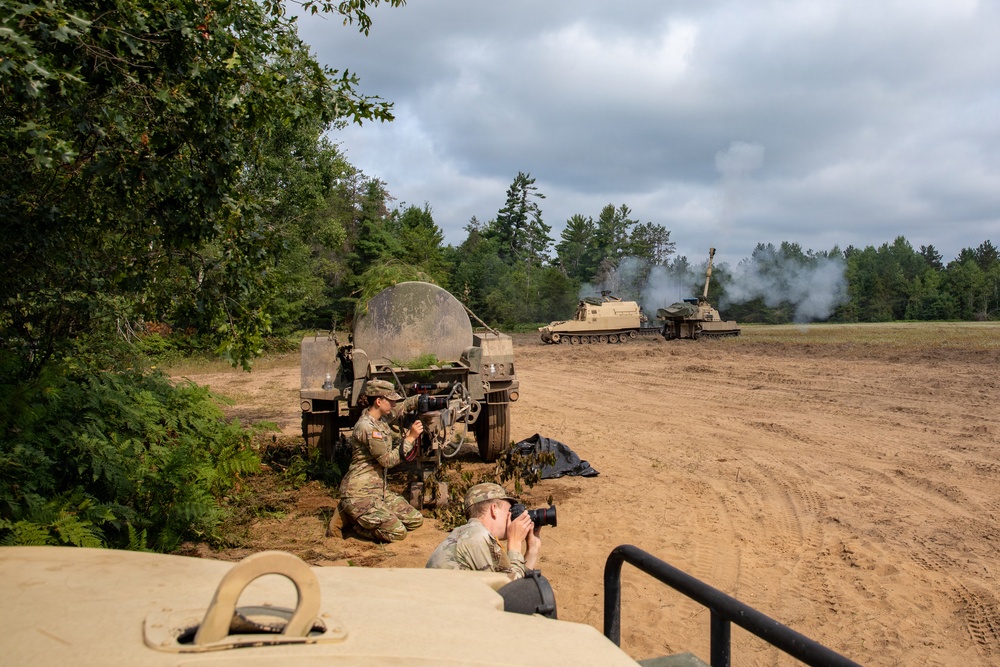 A military training exercise is underway at what appears to be a sandy field range surrounded by forest. Two soldiers in camouflage uniforms are positioned near a military vehicle in the foreground, with one operating equipment and another photographing with a DSLR camera. In the background, a self-propelled howitzer (likely an M109 Paladin) has just fired, with visible smoke rising from its barrel.
