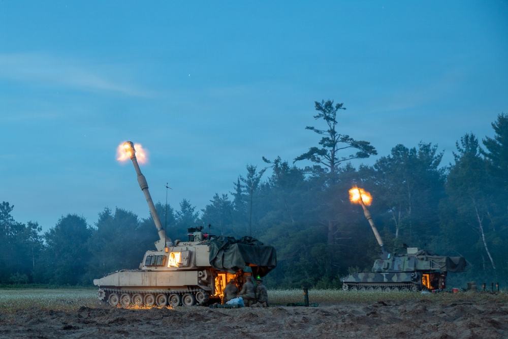 Two self-propelled howitzers are firing simultaneously during twilight or early dawn conditions in an open field near a treeline. Both artillery pieces show muzzle flash at the barrel tips, indicating live fire exercise or combat operation. At least one crew member is visible near the foreground vehicle.