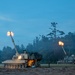 Two self-propelled howitzers are firing simultaneously during twilight or early dawn conditions in an open field near a treeline. Both artillery pieces show muzzle flash at the barrel tips, indicating live fire exercise or combat operation. At least one crew member is visible near the foreground vehicle.