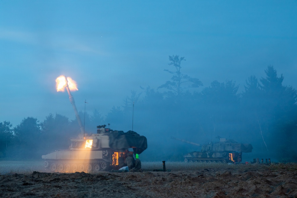 A nighttime or dusk military artillery exercise showing at least two self-propelled howitzers firing in an open field. The foreground vehicle is actively firing, producing a large muzzle flash and illuminating the surrounding smoke and haze. A second artillery vehicle is visible in the background, also positioned for firing.