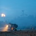 A nighttime or dusk military artillery exercise showing at least two self-propelled howitzers firing in an open field. The foreground vehicle is actively firing, producing a large muzzle flash and illuminating the surrounding smoke and haze. A second artillery vehicle is visible in the background, also positioned for firing.