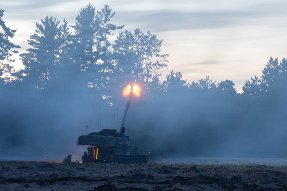 A self-propelled artillery system is captured mid-fire at dusk or dawn, with a bright muzzle flash visible at the end of the elevated barrel. Dense propellant smoke and discharge gases surround the vehicle, partially obscuring the scene. At least one or two personnel are visible near the base of the vehicle on the ground.