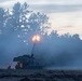 A self-propelled artillery system is captured mid-fire at dusk or dawn, with a bright muzzle flash visible at the end of the elevated barrel. Dense propellant smoke and discharge gases surround the vehicle, partially obscuring the scene. At least one or two personnel are visible near the base of the vehicle on the ground.