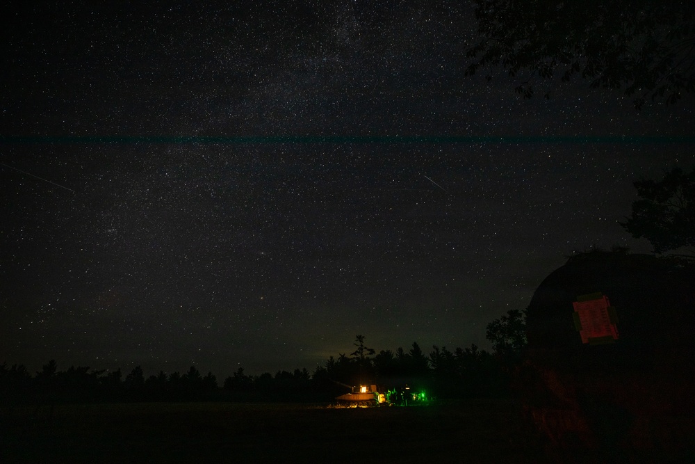 A long-exposure night photography image capturing a star-filled sky with the Milky Way visible. In the foreground, a rural landscape features a tree line, a lit structure with warm orange and green lights, and a dark barn-like building on the right. A faint horizontal teal/cyan line is visible across the upper portion of the sky, likely an atmospheric phenomenon or airglow.