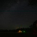 A long-exposure night photography image capturing a star-filled sky with the Milky Way visible. In the foreground, a rural landscape features a tree line, a lit structure with warm orange and green lights, and a dark barn-like building on the right. A faint horizontal teal/cyan line is visible across the upper portion of the sky, likely an atmospheric phenomenon or airglow.