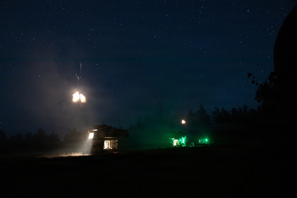 A nighttime military exercise or operation captured in a long-exposure photograph showing two armored vehicles firing their weapons. Muzzle flashes are visible above both vehicles, with one vehicle illuminated by a bright white interior light and the other by green operational lighting. The scene is set against a clear, star-filled night sky with a treeline silhouetted in the background.