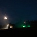 A nighttime military exercise or operation captured in a long-exposure photograph showing two armored vehicles firing their weapons. Muzzle flashes are visible above both vehicles, with one vehicle illuminated by a bright white interior light and the other by green operational lighting. The scene is set against a clear, star-filled night sky with a treeline silhouetted in the background.