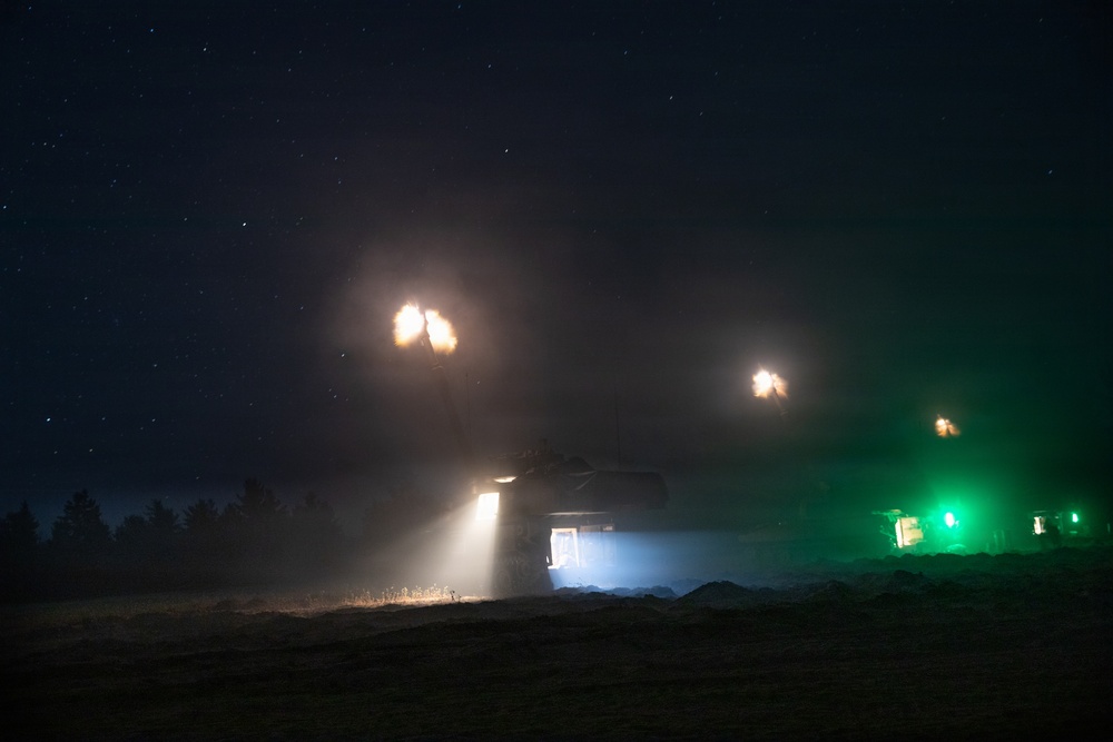 A nighttime military exercise or engagement showing at least one armored vehicle firing its main gun, producing visible muzzle flash and smoke. The scene is illuminated by muzzle blasts, a white spotlight beam from the vehicle, and green marker lights visible on the right side. The starry sky and treeline silhouette are visible in the background.