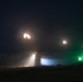 A nighttime military exercise or engagement showing at least one armored vehicle firing its main gun, producing visible muzzle flash and smoke. The scene is illuminated by muzzle blasts, a white spotlight beam from the vehicle, and green marker lights visible on the right side. The starry sky and treeline silhouette are visible in the background.