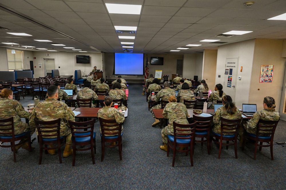 A military training or briefing session is taking place in a conference/classroom setting. Approximately 20-25 uniformed personnel in OCP (Operational Camouflage Pattern) uniforms are seated at tables facing a presenter at a podium. A projection screen displaying a blue slide is visible at the front of the room.