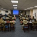 A military training or briefing session is taking place in a conference/classroom setting. Approximately 20-25 uniformed personnel in OCP (Operational Camouflage Pattern) uniforms are seated at tables facing a presenter at a podium. A projection screen displaying a blue slide is visible at the front of the room.