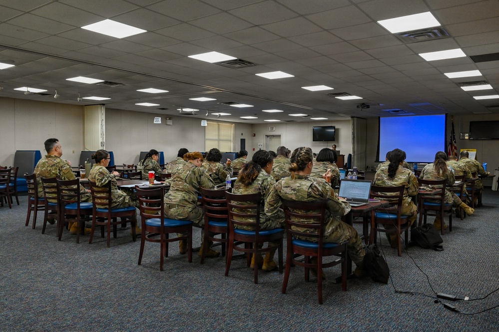 A group of military personnel in camouflage uniforms (OCP/MultiCam pattern) are seated at tables in a conference or briefing room. They appear to be attending a training session or briefing, facing toward a projection screen displaying a blue slide at the front of the room. The setting appears to be a military installation's conference facility.