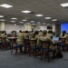 A group of military personnel in camouflage uniforms (OCP/MultiCam pattern) are seated at tables in a conference or briefing room. They appear to be attending a training session or briefing, facing toward a projection screen displaying a blue slide at the front of the room. The setting appears to be a military installation's conference facility.