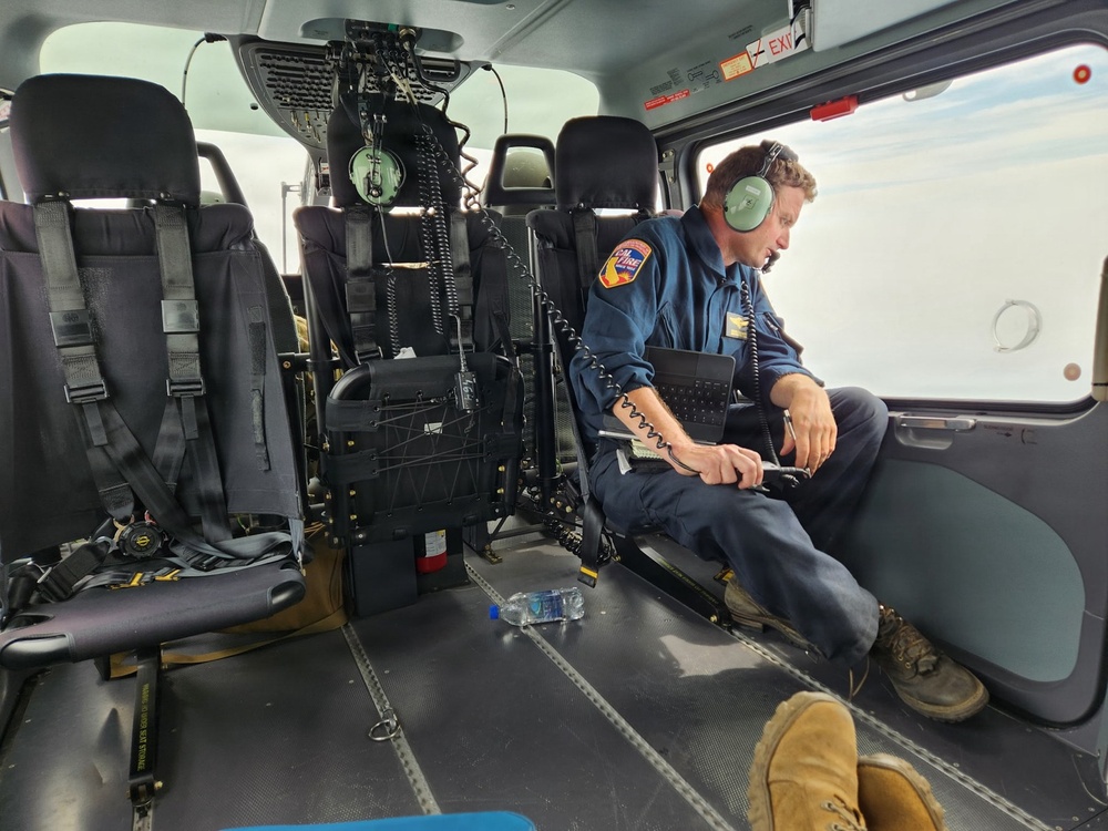 A man in a blue uniform sits inside the cabin of a helicopter, wearing green aviation headphones and holding a radio handset connected by a coiled cord. He appears to be communicating or monitoring radio traffic while in flight, as indicated by the sky visible through the window. His uniform patch suggests affiliation with a fire agency, likely CAL FIRE (California Department of Forestry and Fire Protection).