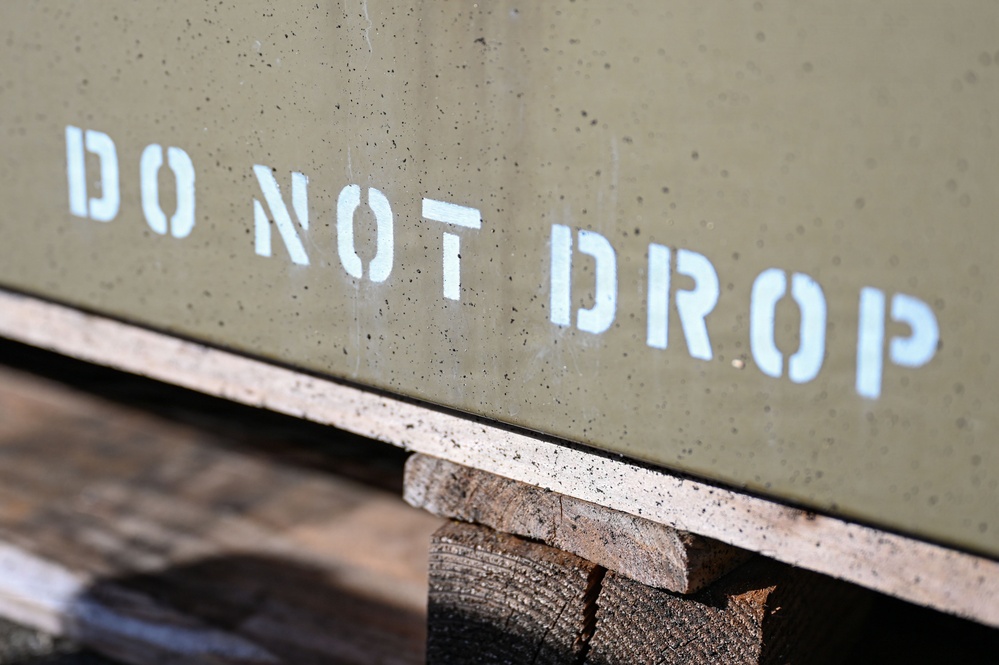 The image shows a close-up of an olive drab military-style metal container or crate with the stenciled warning 'DO NOT DROP' in white block letters. The container is resting on wooden planks or dunnage, typical of military or industrial storage and transport practices. The stenciled text is the primary focal point of the photograph.