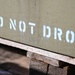 The image shows a close-up of an olive drab military-style metal container or crate with the stenciled warning 'DO NOT DROP' in white block letters. The container is resting on wooden planks or dunnage, typical of military or industrial storage and transport practices. The stenciled text is the primary focal point of the photograph.