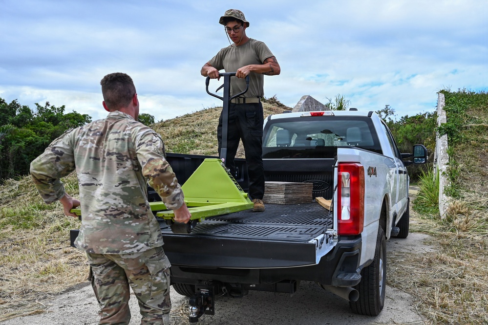 Two individuals are working together to unload or load materials from the bed of a white 4x4 pickup truck in an outdoor, rural setting. One person wearing military camouflage uniform is standing at the tailgate, while another in civilian-style clothing stands in the truck bed operating a bright green pallet jack. A stack of flat materials, possibly tiles or boards, is visible in the truck bed.