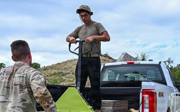 Two individuals are working together to unload or load materials from the bed of a white 4x4 pickup truck in an outdoor, rural setting. One person wearing military camouflage uniform is standing at the tailgate, while another in civilian-style clothing stands in the truck bed operating a bright green pallet jack. A stack of flat materials, possibly tiles or boards, is visible in the truck bed.