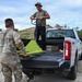 Two individuals are working together to unload or load materials from the bed of a white 4x4 pickup truck in an outdoor, rural setting. One person wearing military camouflage uniform is standing at the tailgate, while another in civilian-style clothing stands in the truck bed operating a bright green pallet jack. A stack of flat materials, possibly tiles or boards, is visible in the truck bed.
