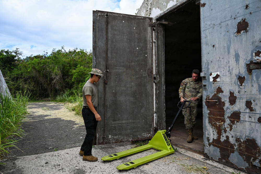 Two military personnel are working at the entrance of an old, heavily weathered concrete bunker or storage structure. One soldier in full camouflage uniform is maneuvering a bright yellow-green pallet jack through the open heavy metal door, while a second person in partial military attire observes. The structure shows significant rust and deterioration on its large metal doors.