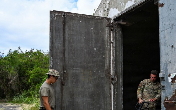 Two military personnel are working at the entrance of an old, heavily weathered concrete bunker or storage structure. One soldier in full camouflage uniform is maneuvering a bright yellow-green pallet jack through the open heavy metal door, while a second person in partial military attire observes. The structure shows significant rust and deterioration on its large metal doors.