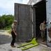 Two military personnel are working at the entrance of an old, heavily weathered concrete bunker or storage structure. One soldier in full camouflage uniform is maneuvering a bright yellow-green pallet jack through the open heavy metal door, while a second person in partial military attire observes. The structure shows significant rust and deterioration on its large metal doors.