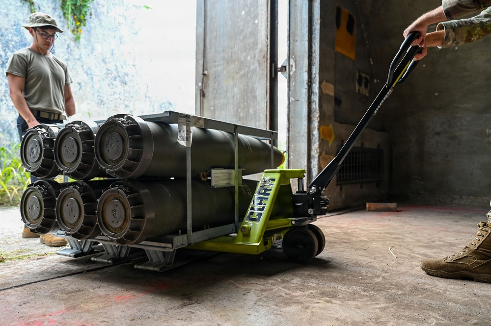 Two military personnel in camouflage/olive drab uniforms are handling a rack of what appear to be munitions or ordnance cylinders loaded on a bright green CLARK hydraulic pallet jack. The scene takes place at the entrance of what appears to be a concrete storage bunker or ammunition storage facility. One service member is guiding the load from the rear while another observes from the side near the doorway.