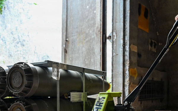 Two military personnel in camouflage/olive drab uniforms are handling a rack of what appear to be munitions or ordnance cylinders loaded on a bright green CLARK hydraulic pallet jack. The scene takes place at the entrance of what appears to be a concrete storage bunker or ammunition storage facility. One service member is guiding the load from the rear while another observes from the side near the doorway.
