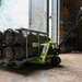 Two military personnel in camouflage/olive drab uniforms are handling a rack of what appear to be munitions or ordnance cylinders loaded on a bright green CLARK hydraulic pallet jack. The scene takes place at the entrance of what appears to be a concrete storage bunker or ammunition storage facility. One service member is guiding the load from the rear while another observes from the side near the doorway.