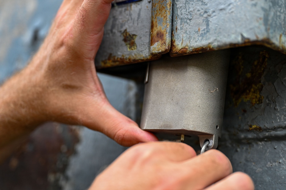 A person is using both hands to manipulate or install a metal component on a heavily rusted metal surface. The new or cleaner metal piece appears to be a bracket, cover, or housing being fitted into an existing corroded metal structure. The activity suggests maintenance, repair, or installation work on aging metal equipment or infrastructure.