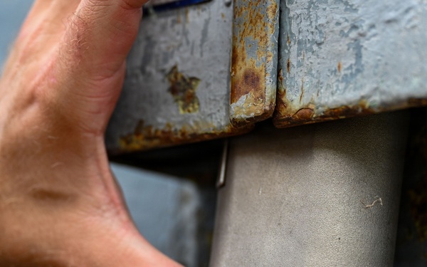 A person is using both hands to manipulate or install a metal component on a heavily rusted metal surface. The new or cleaner metal piece appears to be a bracket, cover, or housing being fitted into an existing corroded metal structure. The activity suggests maintenance, repair, or installation work on aging metal equipment or infrastructure.