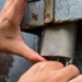 A person is using both hands to manipulate or install a metal component on a heavily rusted metal surface. The new or cleaner metal piece appears to be a bracket, cover, or housing being fitted into an existing corroded metal structure. The activity suggests maintenance, repair, or installation work on aging metal equipment or infrastructure.