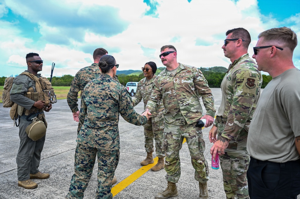 A group of U.S. military personnel from multiple branches are gathered on what appears to be a flight line or tarmac in a tropical setting. A Marine in MARPAT digital camouflage is shaking hands with a U.S. Air Force service member in OCP (Operational Camouflage Pattern) uniform, suggesting an inter-service greeting or coordination meeting. The setting and mix of uniforms indicate a joint military operation or exercise.