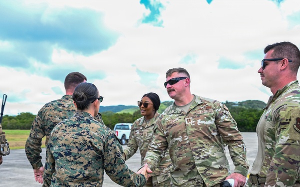 A group of U.S. military personnel from multiple branches are gathered on what appears to be a flight line or tarmac in a tropical setting. A Marine in MARPAT digital camouflage is shaking hands with a U.S. Air Force service member in OCP (Operational Camouflage Pattern) uniform, suggesting an inter-service greeting or coordination meeting. The setting and mix of uniforms indicate a joint military operation or exercise.