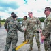 A group of U.S. military personnel from multiple branches are gathered on what appears to be a flight line or tarmac in a tropical setting. A Marine in MARPAT digital camouflage is shaking hands with a U.S. Air Force service member in OCP (Operational Camouflage Pattern) uniform, suggesting an inter-service greeting or coordination meeting. The setting and mix of uniforms indicate a joint military operation or exercise.