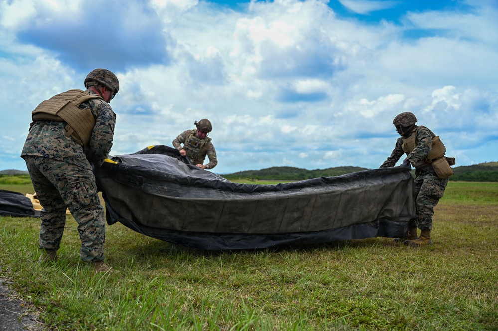Three military personnel in combat uniforms and helmets are working together on an open grassy field to fold or pack a large dark-colored parachute or tarpaulin. The soldiers appear to be conducting post-jump recovery or equipment packing operations. The setting appears to be a tropical or subtropical environment based on the lush green vegetation and cumulus cloud formations.