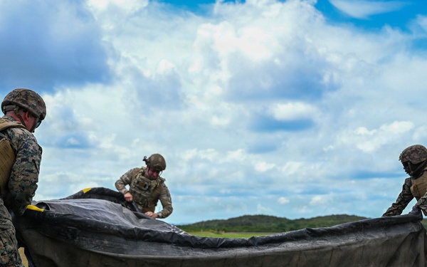 Three military personnel in combat uniforms and helmets are working together on an open grassy field to fold or pack a large dark-colored parachute or tarpaulin. The soldiers appear to be conducting post-jump recovery or equipment packing operations. The setting appears to be a tropical or subtropical environment based on the lush green vegetation and cumulus cloud formations.