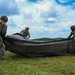 Three military personnel in combat uniforms and helmets are working together on an open grassy field to fold or pack a large dark-colored parachute or tarpaulin. The soldiers appear to be conducting post-jump recovery or equipment packing operations. The setting appears to be a tropical or subtropical environment based on the lush green vegetation and cumulus cloud formations.