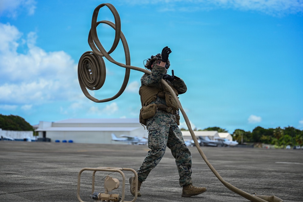 A U.S. Marine in woodland MARPAT camouflage uniform is handling a large fuel or water hose on an airfield tarmac, appearing to coil or deploy it with significant physical effort. The marine is wearing full tactical gear including a plate carrier, black gloves, and hearing protection. The setting appears to be a tropical airfield with civilian aircraft and hangars visible in the background.