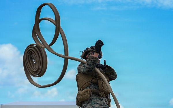 A U.S. Marine in woodland MARPAT camouflage uniform is handling a large fuel or water hose on an airfield tarmac, appearing to coil or deploy it with significant physical effort. The marine is wearing full tactical gear including a plate carrier, black gloves, and hearing protection. The setting appears to be a tropical airfield with civilian aircraft and hangars visible in the background.