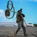 A U.S. Marine in woodland MARPAT camouflage uniform is handling a large fuel or water hose on an airfield tarmac, appearing to coil or deploy it with significant physical effort. The marine is wearing full tactical gear including a plate carrier, black gloves, and hearing protection. The setting appears to be a tropical airfield with civilian aircraft and hangars visible in the background.