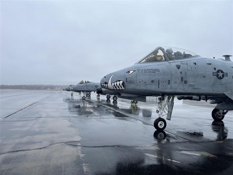 A line of A-10 Thunderbolt II (Warthog) ground-attack aircraft are parked on a wet tarmac under overcast, rainy conditions. The aircraft display the iconic shark-mouth nose art and U.S. Air Force markings. At least one aircraft has a pilot or crew member visible in the cockpit, suggesting pre-flight or ground operations.