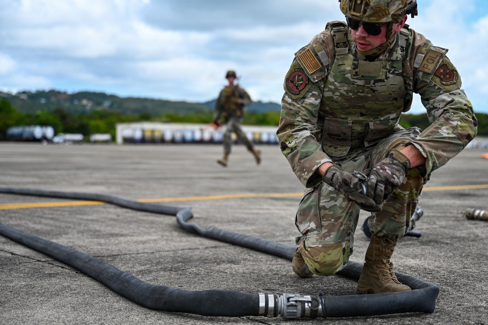 A U.S. Air Force service member in full MultiCam combat gear is crouched on a tarmac handling a large black fuel hose with a metal coupling. A second uniformed service member is visible running in the background. The setting appears to be an airfield, likely during a forward area refueling point (FARP) or petroleum, oil, and lubricants (POL) training exercise.