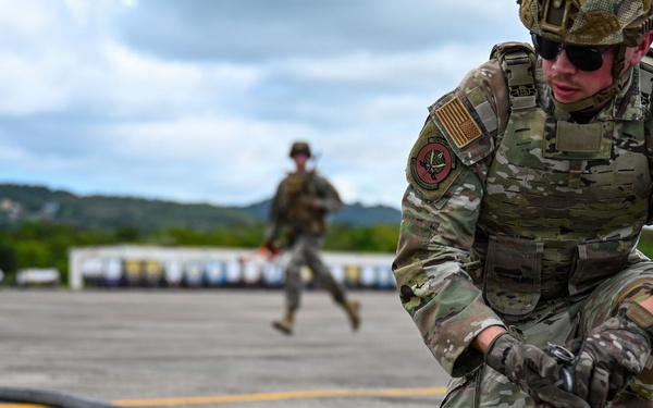 A U.S. Air Force service member in full MultiCam combat gear is crouched on a tarmac handling a large black fuel hose with a metal coupling. A second uniformed service member is visible running in the background. The setting appears to be an airfield, likely during a forward area refueling point (FARP) or petroleum, oil, and lubricants (POL) training exercise.