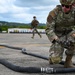 A U.S. Air Force service member in full MultiCam combat gear is crouched on a tarmac handling a large black fuel hose with a metal coupling. A second uniformed service member is visible running in the background. The setting appears to be an airfield, likely during a forward area refueling point (FARP) or petroleum, oil, and lubricants (POL) training exercise.